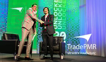 Vlad Tenev in a black suit shakes hands with Robb Baldwin in tan suit on TradePMR Synergy25 conference stage. 